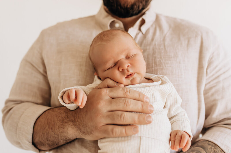 A newborn sleeping peacefully in dad's arms. Dad holds him against his chest with his hands wrapped around him.