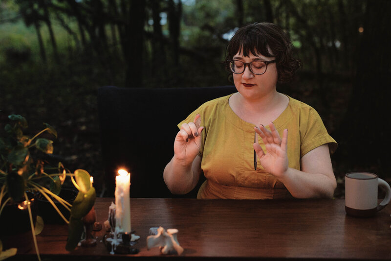 Ash sits at a wood table in the trees practicing energy work wearing a mustard yellow dress