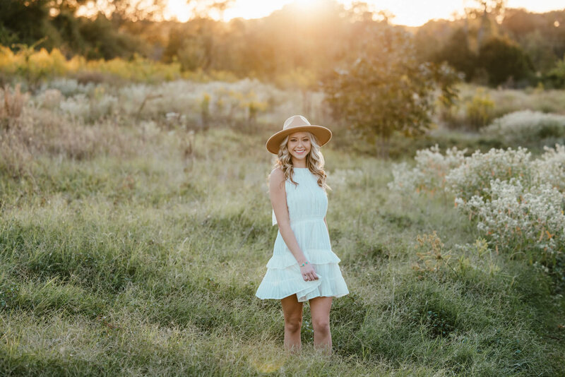 senior girl in short white dress and light brown hat walking through field at sunset
