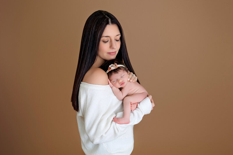 A mother gently cradles her newborn baby against her chest, both looking peaceful and content. The mother has long, straight black hair and is wearing a soft, off-the-shoulder white sweater. The newborn is swaddled in a diaper and has a delicate headband with a floral design. The background is a warm, light brown, adding a cozy and intimate feel to the portrait. The mother's serene expression and the baby's relaxed pose convey a sense of love and tenderness.