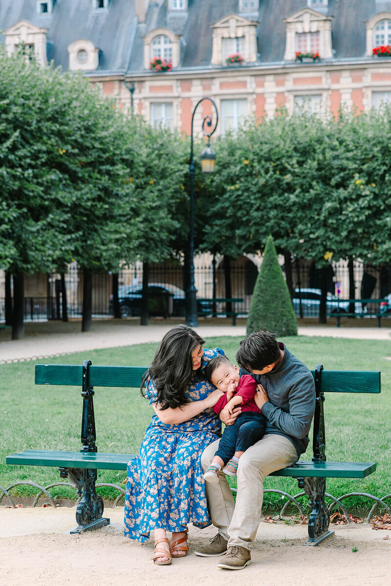 Playful family of three — mother, father, and child — sitting together on a bench during a joyful Paris family photoshoot.