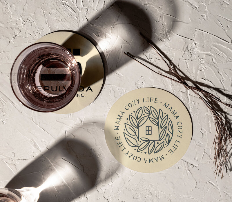 Mama Cozy Life logo printed on a round coaster with an elegant wreath and home icon, representing a warm and inviting brand identity.