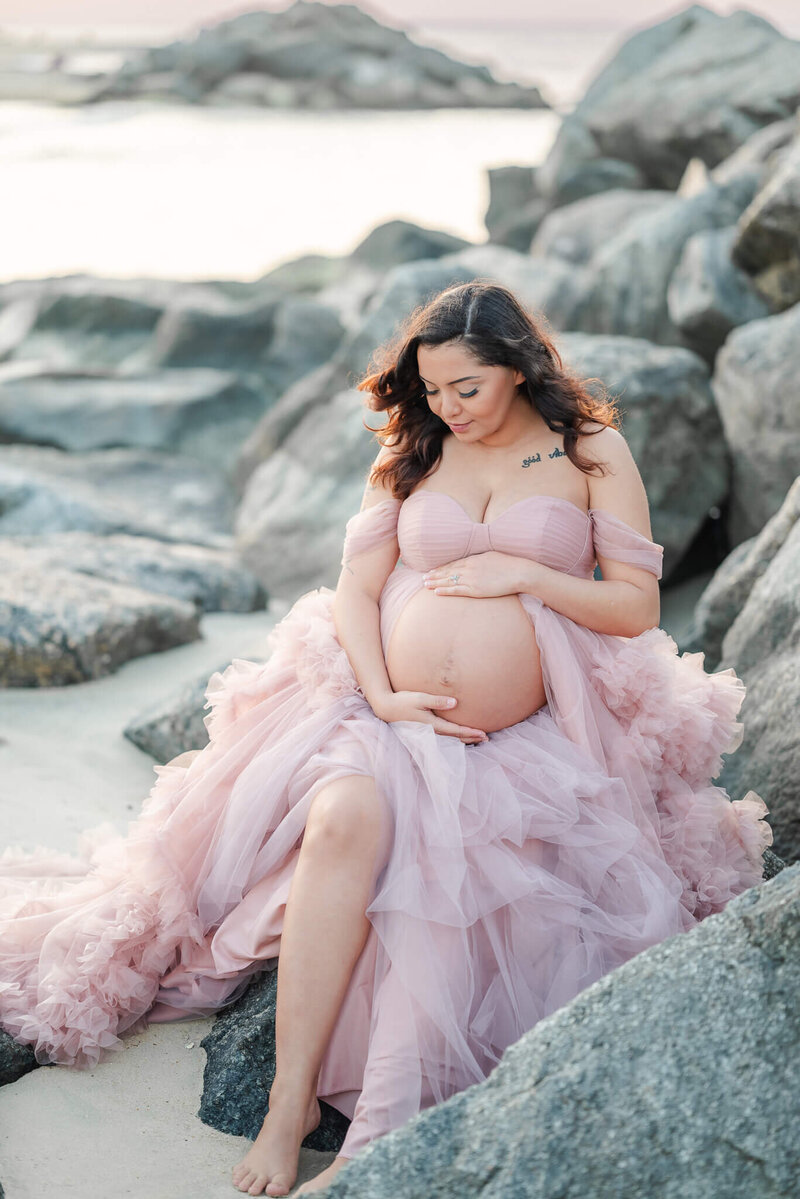 A pregnant woman, wearing a pink tulle gown, looks down at her belly. She sits on some rocks on the beach and cradles her belly.