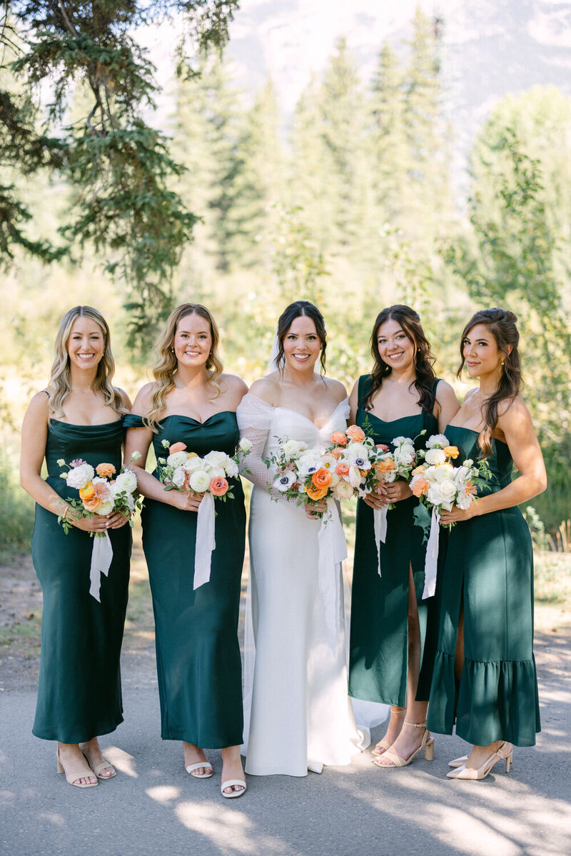 The bride and her bridesmaid stand together outside the Malcolm Hotel in Canmore, Alberta, sharing a joyful moment as they look toward the camera, framed by the stunning mountain backdrop.