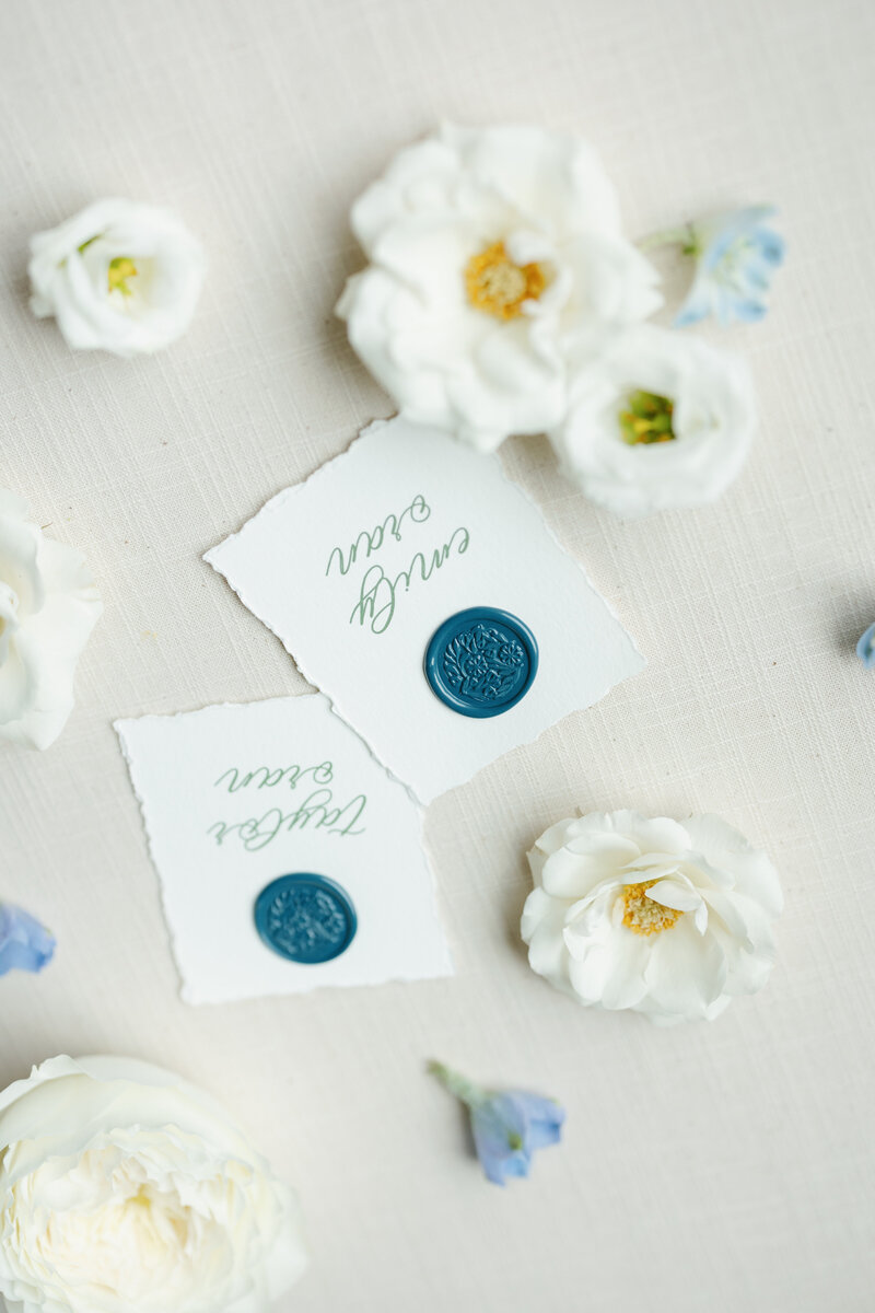 Custom wedding place cards with blue wax seals and hand-lettered calligraphy, styled with white garden roses and soft blue blooms by Eventive Designs.