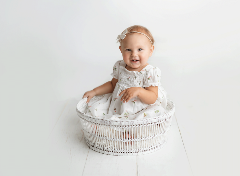 A toddler baby girl in a white dress sits in a wicker basket in a studio