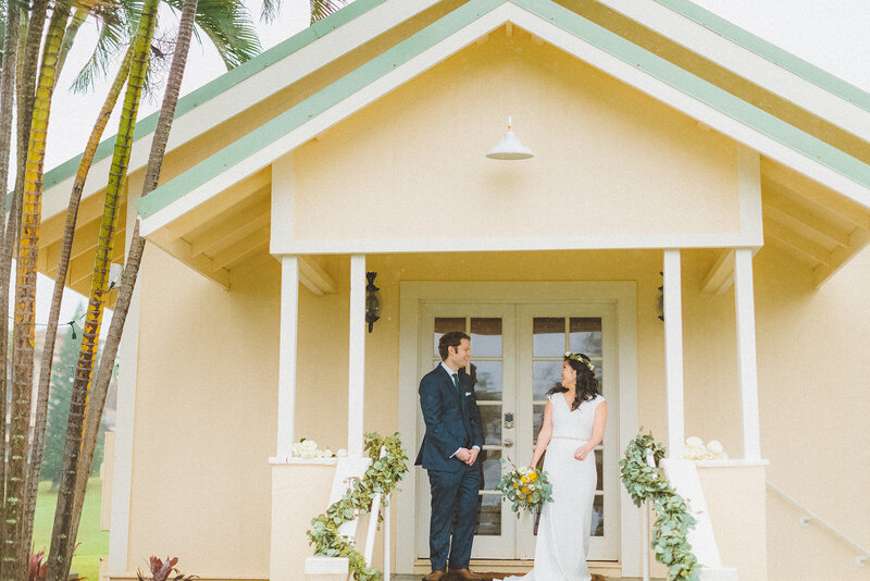 newly married bride and groom pose on the front steps of an old yellow chapel adorned with greenery along the staircase