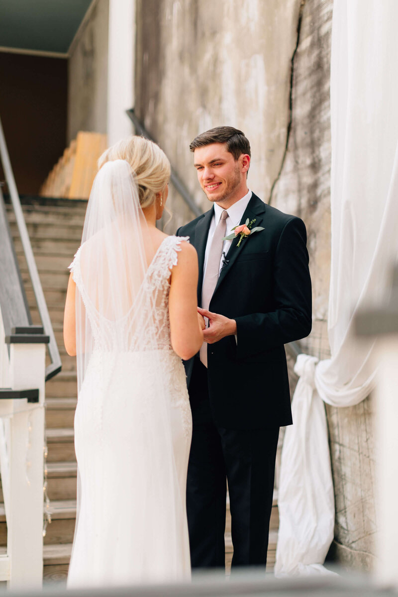 A bride in a white dress and veil smiles at a groom in a black suit and tie. They stand next to a rustic brick wall and wooden stairs, sharing a joyful moment together.