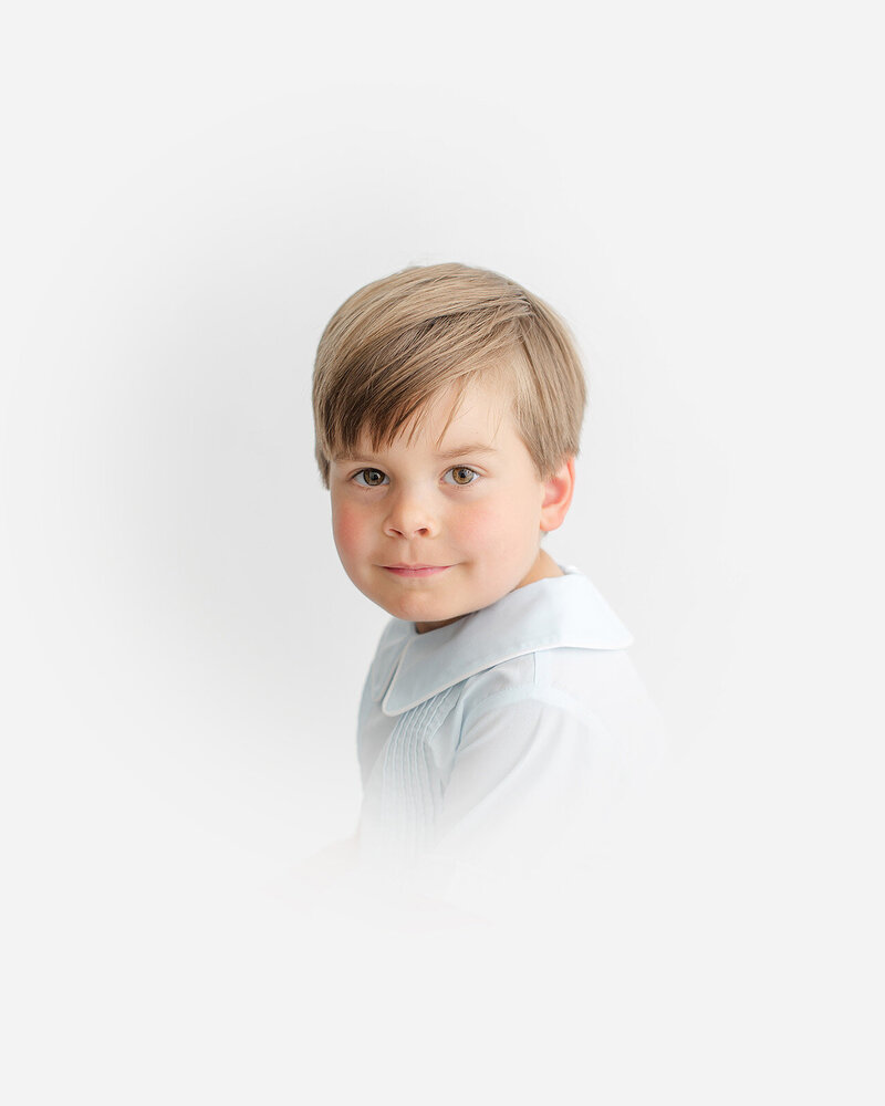 A young child with light brown hair, wearing a light blue collared shirt, is captured in this charming portrait photography against a crisp white background.