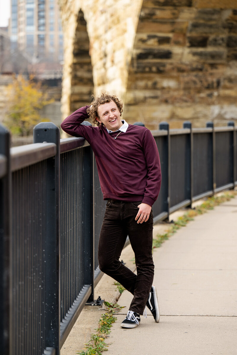 senior photo of guy wearing maroon sweater and dark jeans leaning against a fence