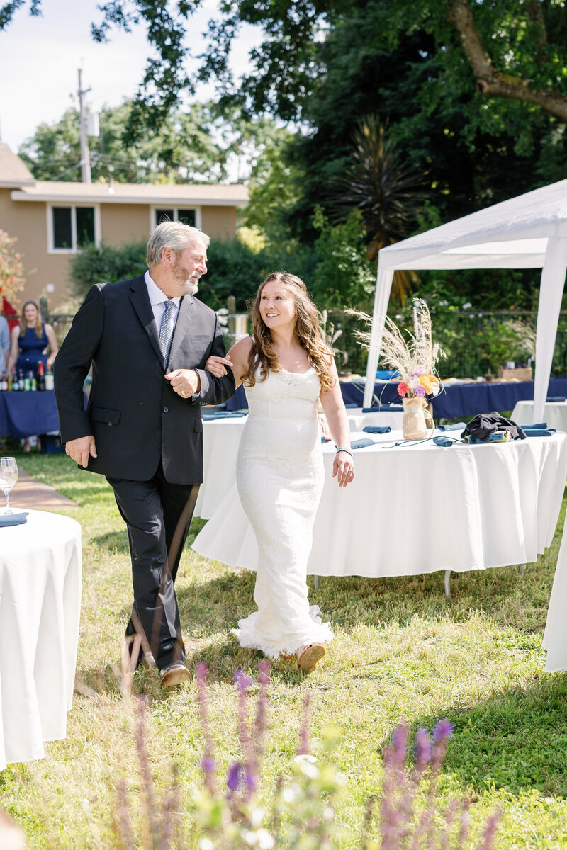 bay area photographers bride and her father walking together into the wedding venue locked in arms past white tables with blue decor accents for a California wedding