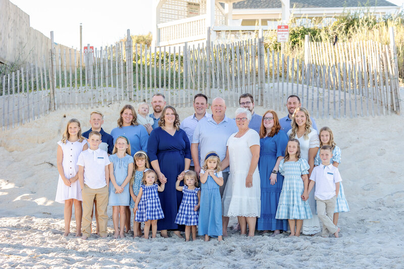 A large family stands together on sandy dunes with a beach house in the background, dressed in coordinating blue and white outfits. The scene captures a classic coastal family moment, ideal for destination family photographer NC, showcasing unity and elegance.