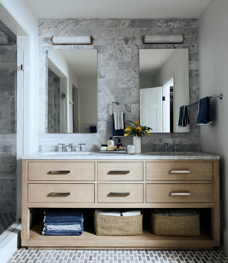 A modern primary bathroom in a historic Hingham home by the best Boston interior designers, featuring an oak vanity, marble backsplash, mosaic flooring, and polished nickel fixtures.