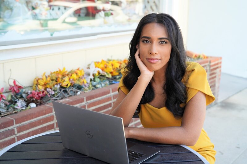 Tania in a yellow dress on laptop looking at camera, with flowers and a brick wall behind her
