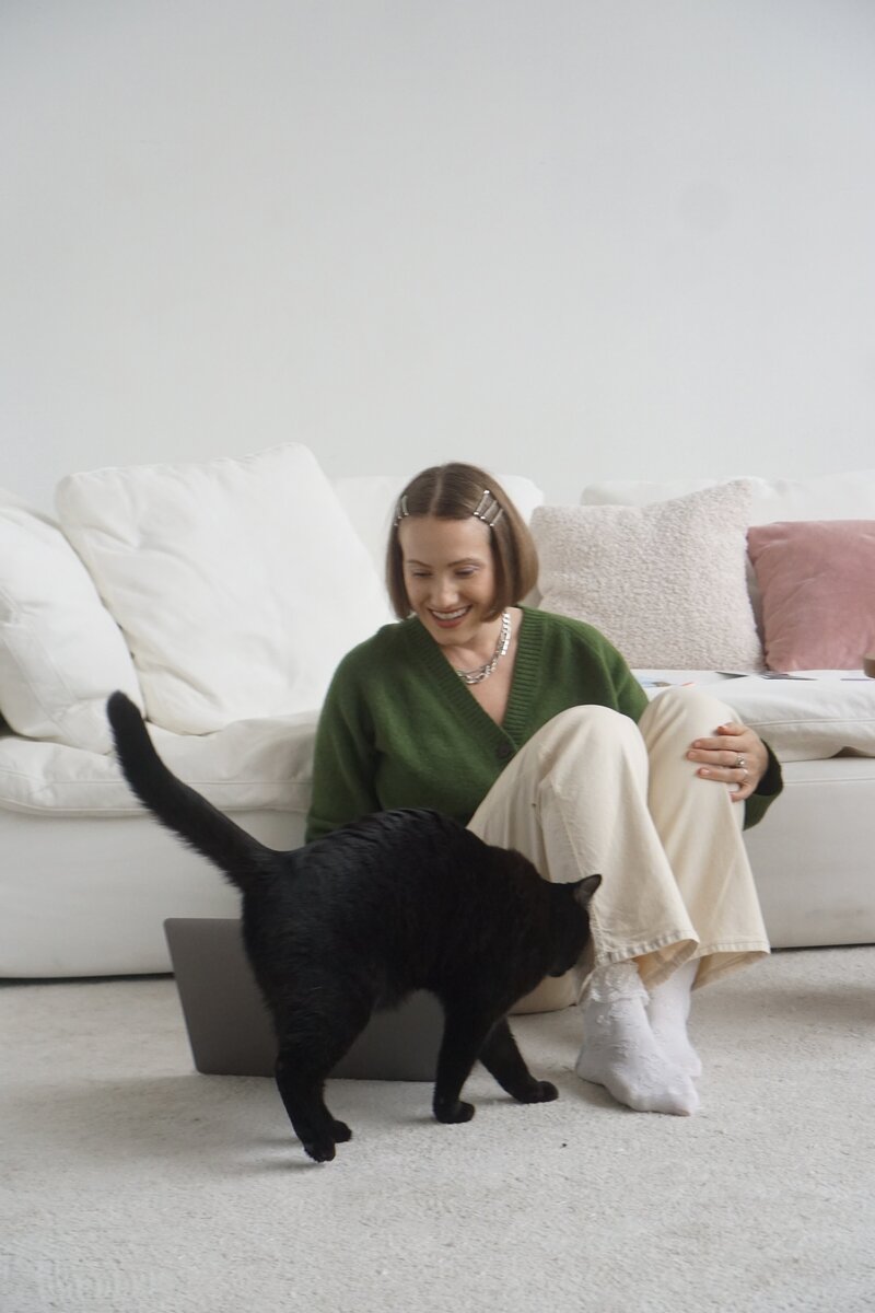 Margarita sits on a carpeted floor,  smiling and looking at her laptop while a  black cat passes by in front.
