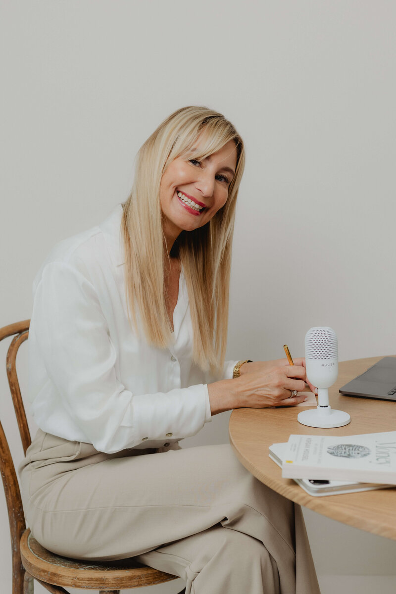 AA women sitting at a table smiling with a microphone and a pen in her hand.
