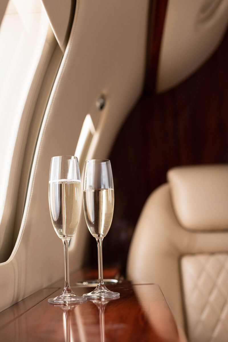 Two glasses of champagne on a polished wooden table inside a luxurious private jet with beige leather seats.