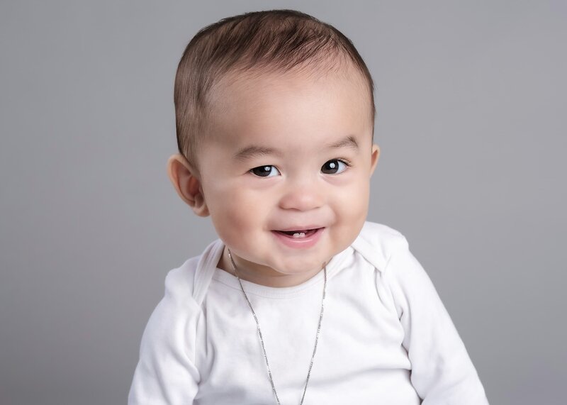 A baby boy in a white onesie, wearing a simple silver chain, smiling brightly with a soft gray background for his first birthday portrait.