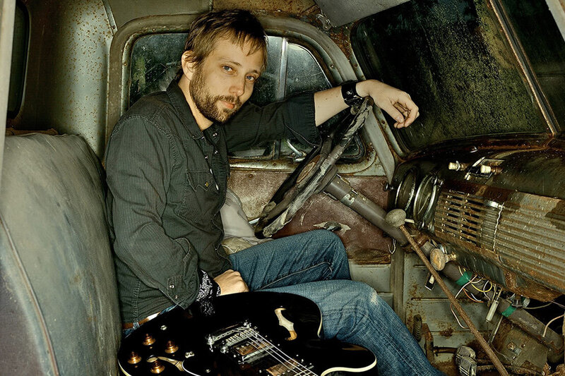 Musician Portrait Ryan McMahon sitting behind wheel of old truck black electric guitar on seat beside him