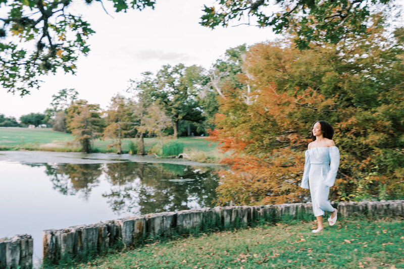 Woman in a blue dress walking beside water in the grass with trees behind her