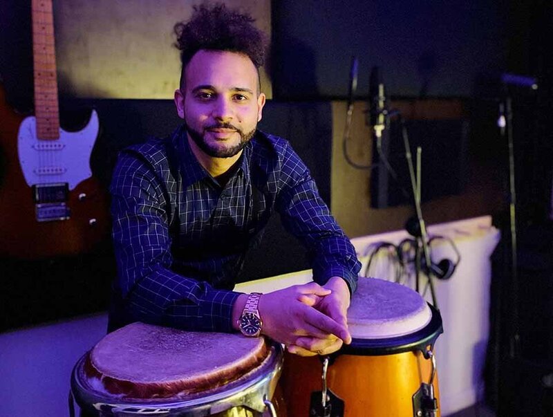Percussionist leaning on conga drums inside a studio setup with guitar and microphones in the background.