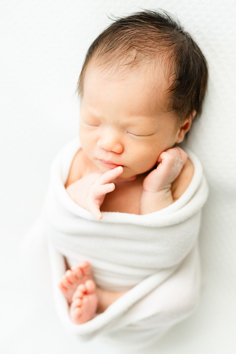 a sleeping infant girl wrapped in a white baby swaddle lays on a white bean bag for her newborn photos.