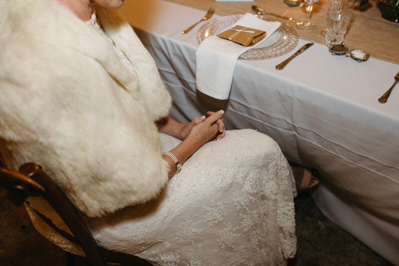 A bride sits and listens to speeches at her reception , there are table decoration details and her dress details can be seen