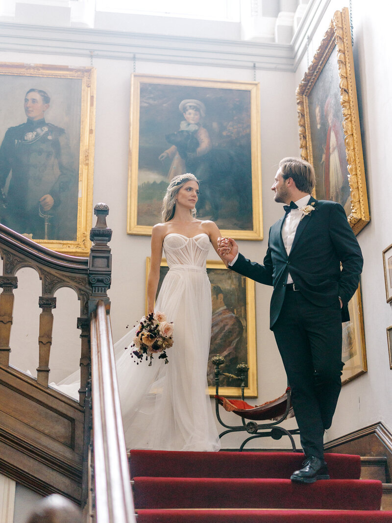 Groom leading bride down a large staircase with red carpet and ornate gold framed paintings on the walls