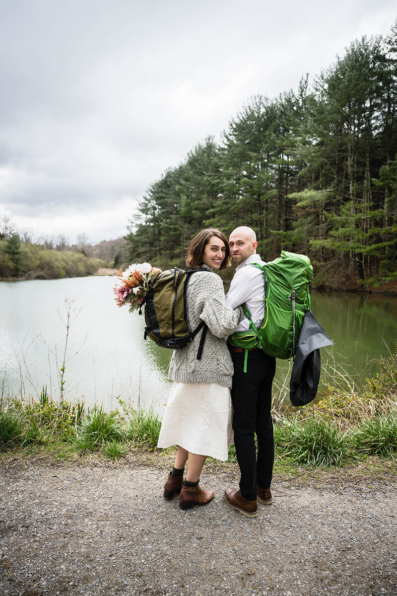 A bride and groom wearing hiking backpacks stand next to one another and look back for a photo. The pair stands in front of a mill pond at Heritage Park.
