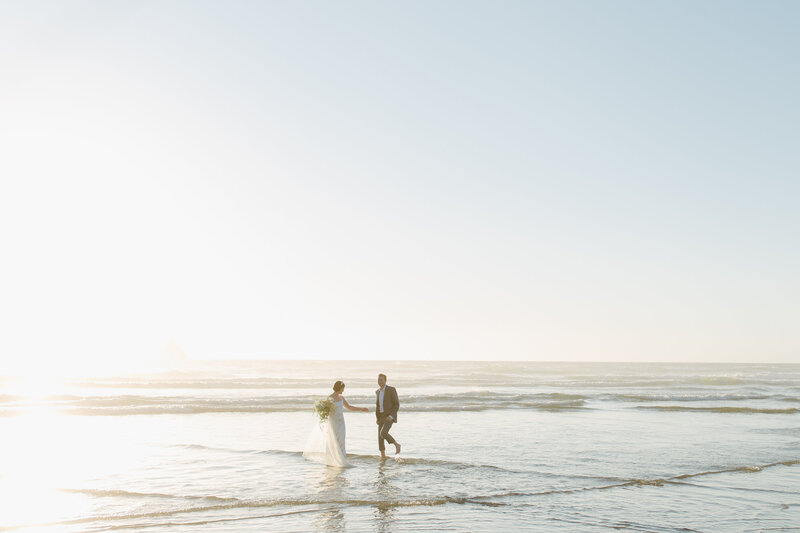 Landscape shot of bride and groom with feet in water at sunset  at Arch Cape in Oregon.