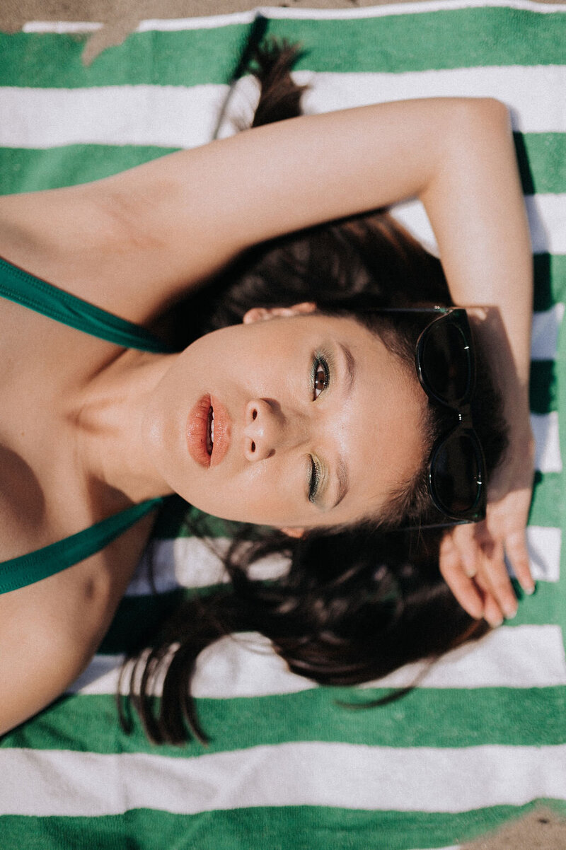 Woman winks wearing 50's style green swimsuit laying on a green and white striped towel with arm bent behind her head.