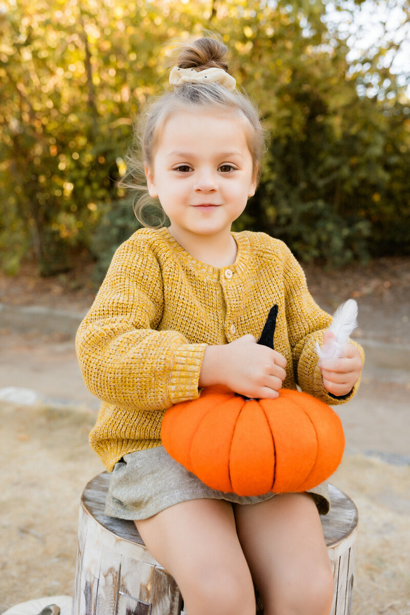 Young girl wearing sweater sits on a log holding a pumpkin