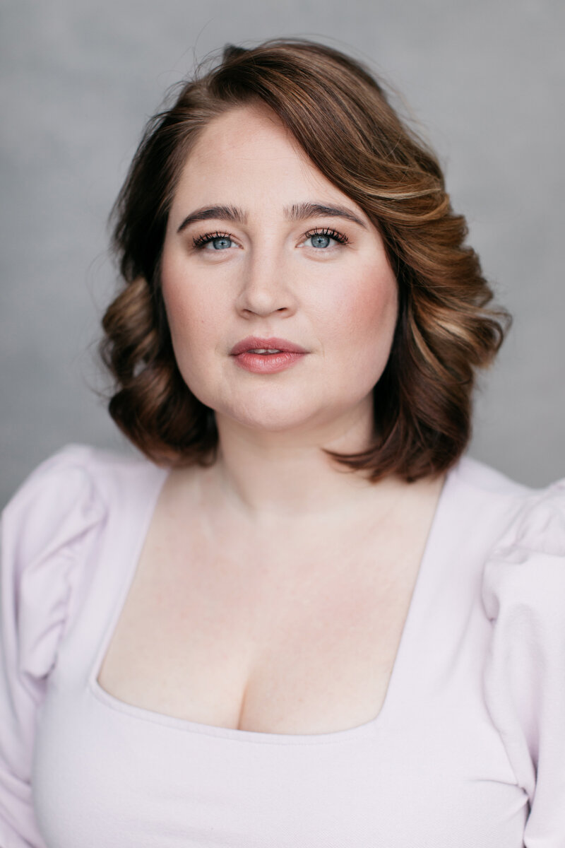 Casting headshot van actrice met bruin haar in lichtroze top voor grijze backdrop.