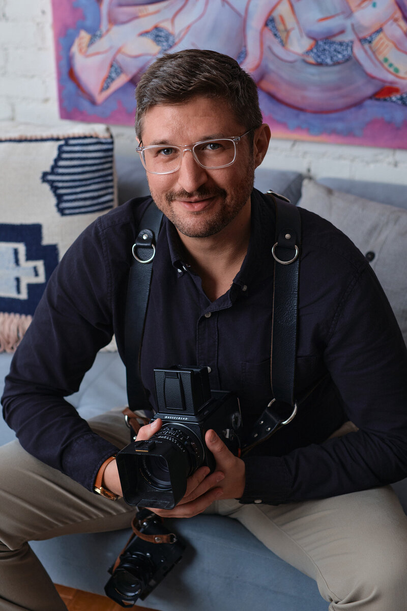 A man with glasses and a short beard, dressed in a dark shirt and tan pants, sits on a couch holding a camera. As an NJ wedding photographer, he wears a camera harness and smiles at the camera. Colorful artwork and cushions are visible in the background.
