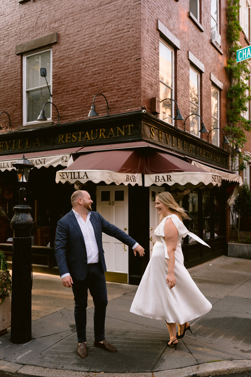 an engaged couple standing next to each other while laughing. Man is reaching out his hand towards the bride-to-be while she is twirling in a white dress
