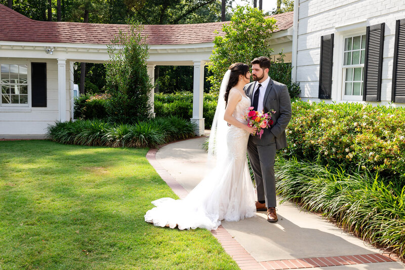 Bride and Groom during golden hour, sunset photos on hill of a golf course club wedding venue with a pavilion in the background.  Orange, gray and blue skies with dramatic cloud and sun peeking through.  Bride in an off the shoulder gown with a long veil.  Groom in a tuxedo with black pants, white jacket and forest green vest.  Smiling and embracing each other.  Virginia Wedding Photography by Angela Foushee at The Venue at Cahoon in Chesapeake VA