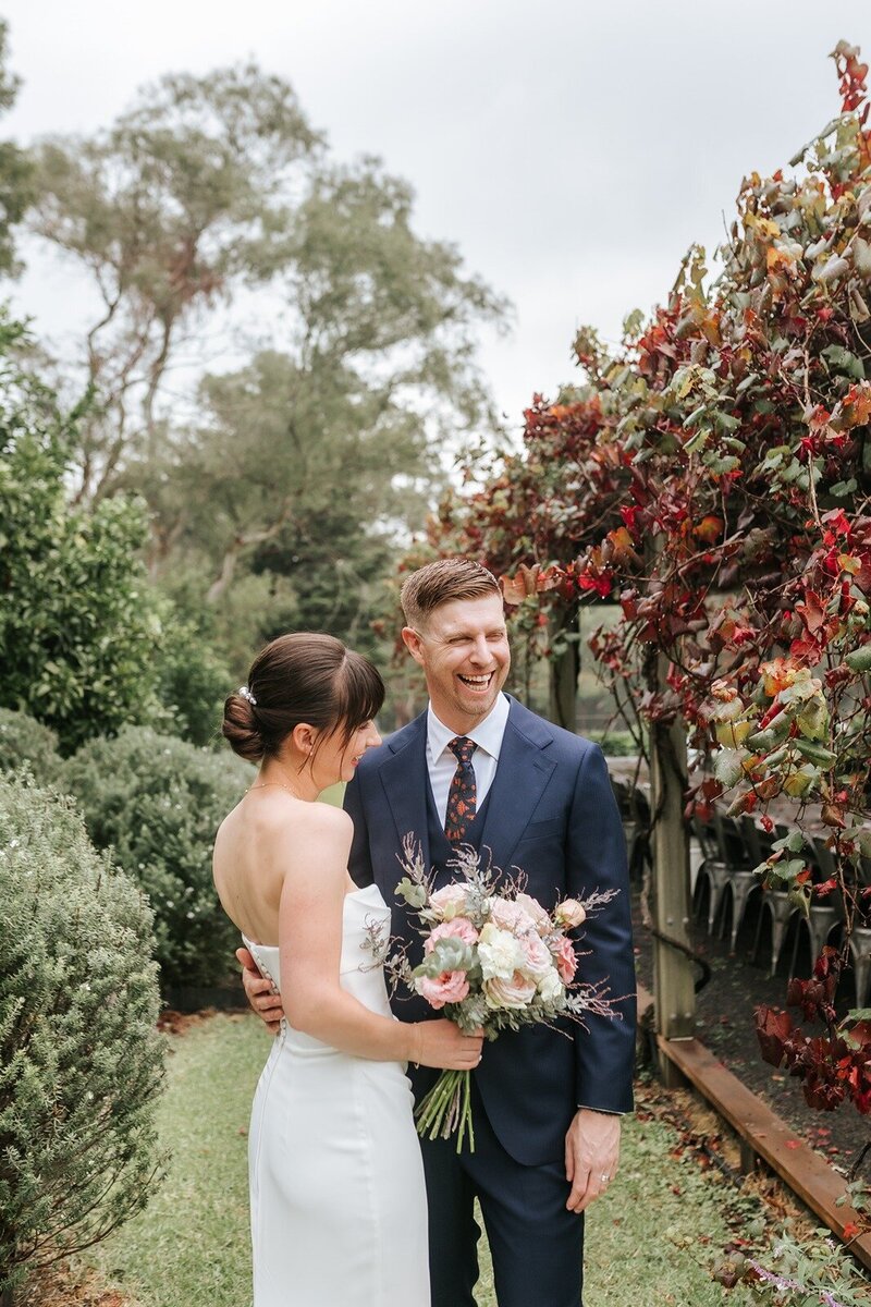 A laughing groom standing with his bride in pretty gardens