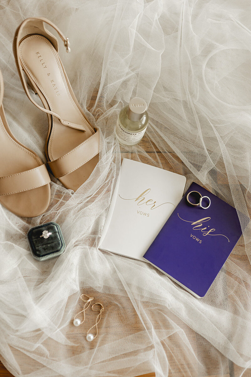 A flat-lay photo of wedding details including beige sandals, a bottle of perfume, wedding rings, a ‘his’ and ‘her’ vow book, pearl earrings, and a jewelry box. The items are arranged on a layer of tulle fabric.