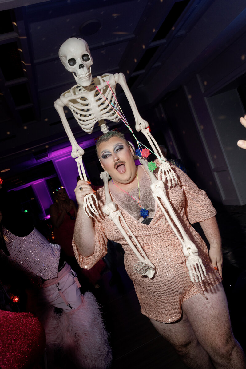 wedding guest in drag on the dance floor with a skeleton on their shoulders looking excited