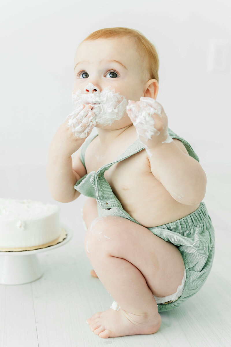 Baby in green romper looking up while eating cake during photography session in Dothan Alabama studio