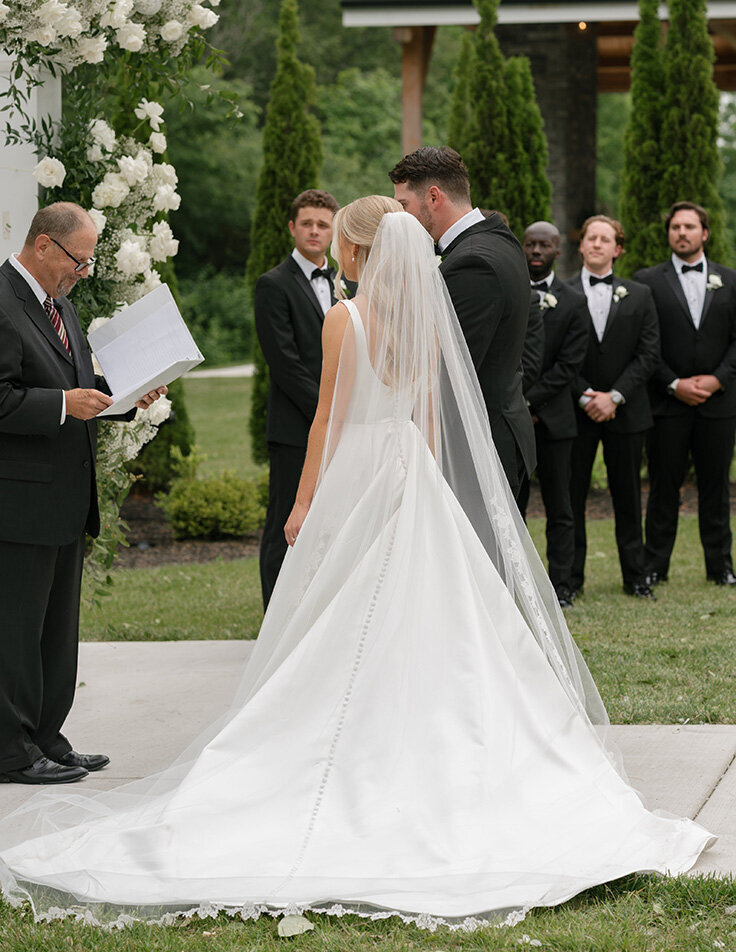 A Columbus bride in a flowing white gown and veil stands next to the groom at an outdoor wedding ceremony. The officiant reads from a booklet, and groomsmen in black suits watch solemnly.