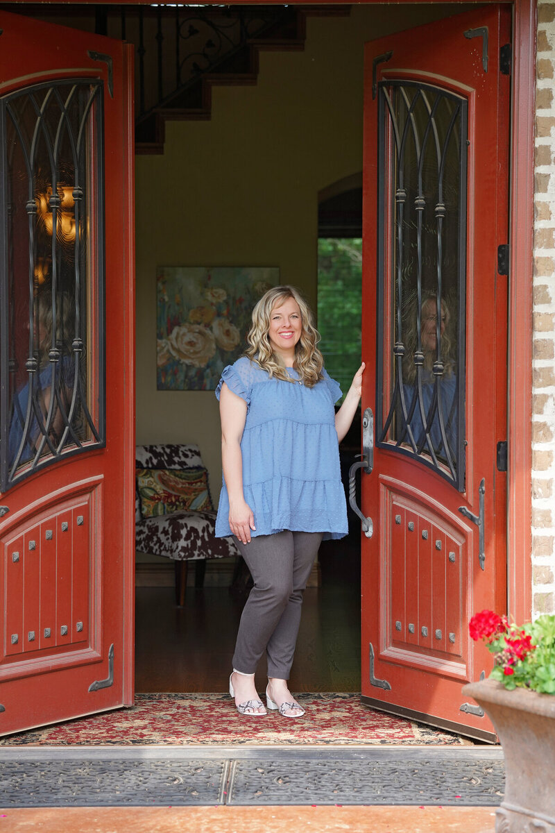 A woman stands in the doorway of a house with red double doors, wearing a blue blouse and grey pants, inviting visitors in to view her Showit web design for personal brands.