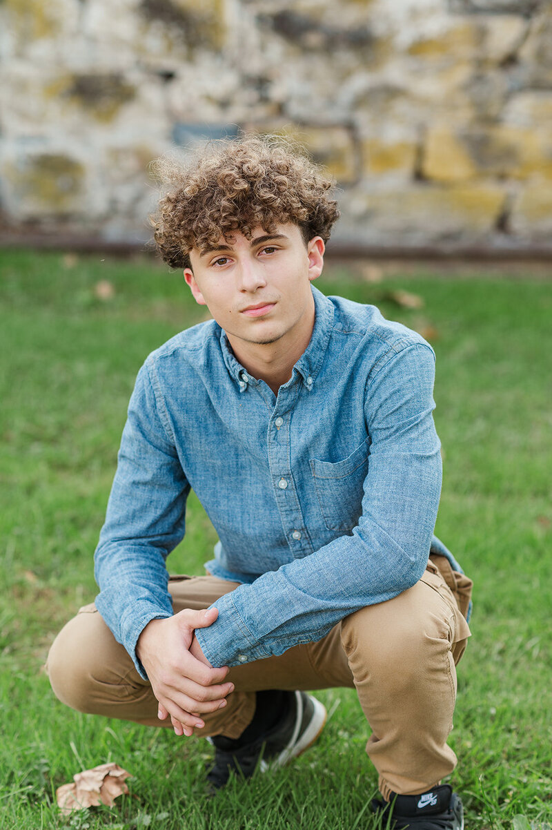 Senior boy, wearing a blue button down shirt and brown pants kneeling in the grass by Lancaster PA photographer