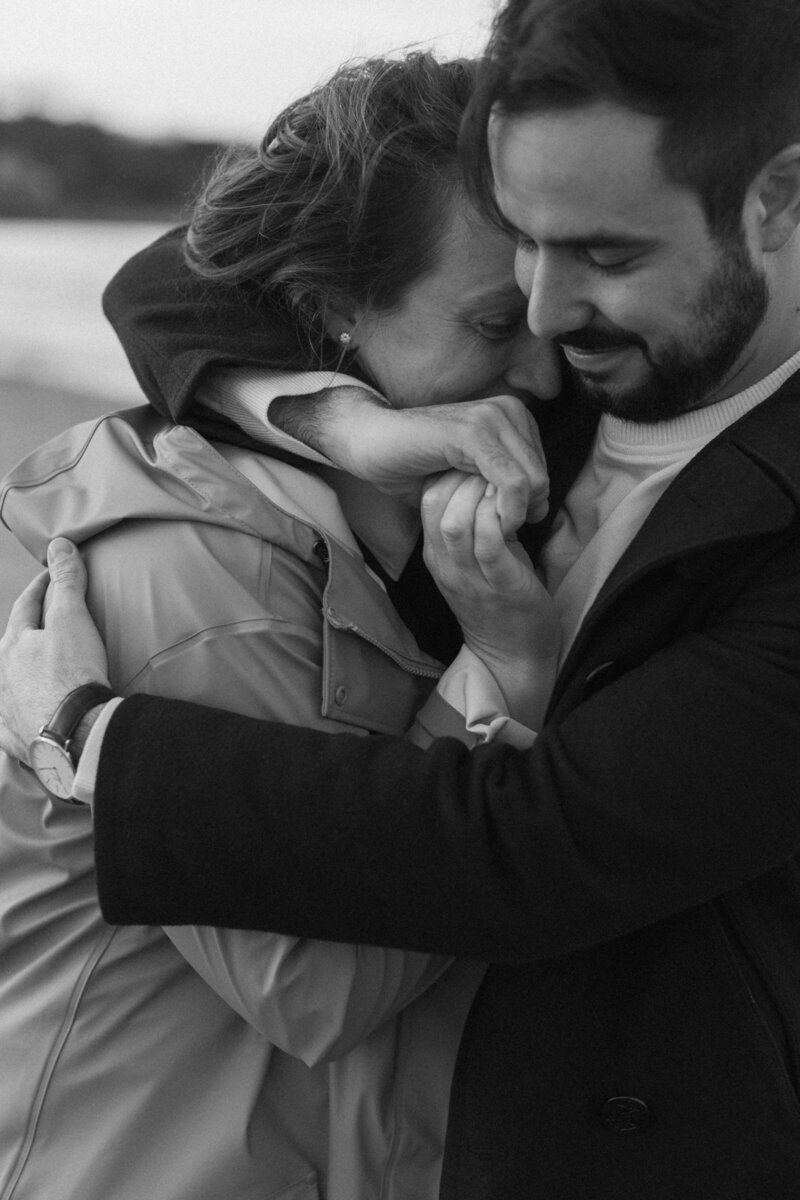 An engaged couple cuddles closely in black and white on a beach in maine