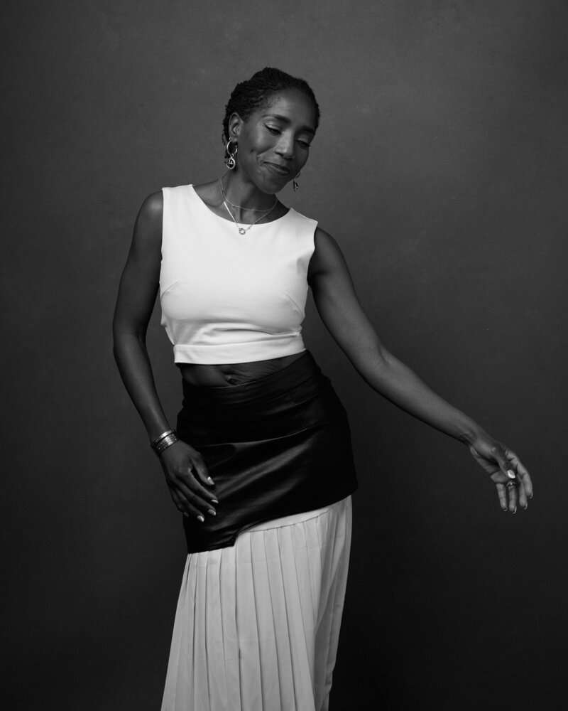 Black and white portrait of a woman moving gracefully, eyes closed with a serene smile. She wears a cropped white top, a black asymmetrical leather skirt over a flowing pleated skirt, and delicate jewelry. Her arm extends softly, exuding elegance, confidence, and self-expression. A beautiful, unfiltered moment from 'THIS IS ME – A Photography Experience by Katie Campbell' in Atlanta, GA, celebrating authenticity and movement.
