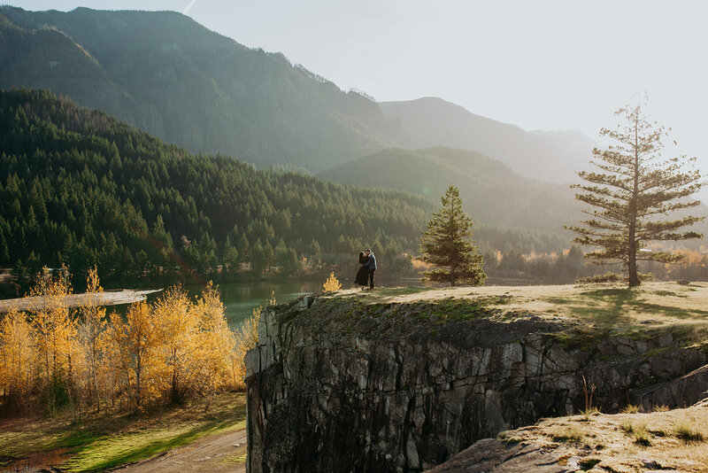 two couple stand on cliff embracing with forest in background