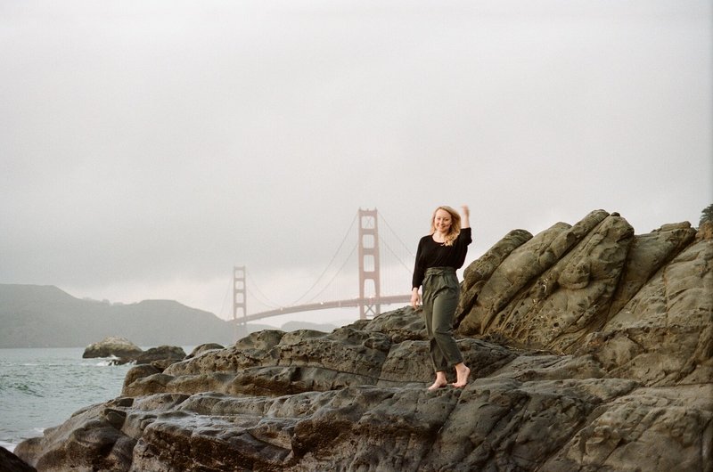 Bridget stands on rocks with the Golden Gate Bridge behind her in San Francisco