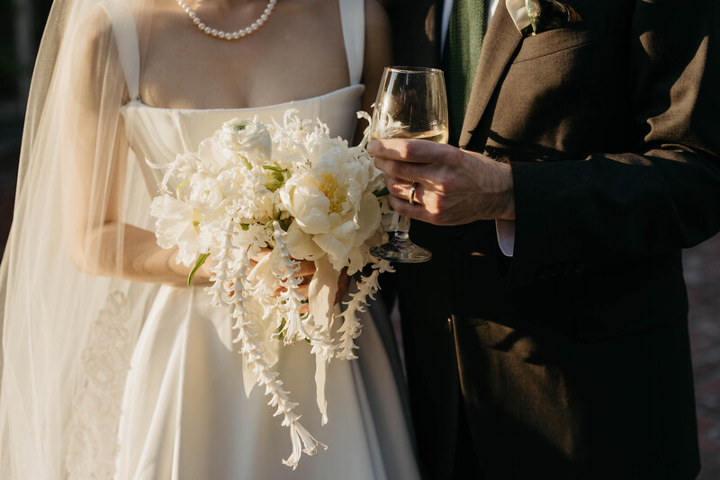 A close-up of a bride holding a bouquet of white flowers and a wine glass, dressed in a simple, elegant gown with a pearl necklace, standing next to the groom in a suit.