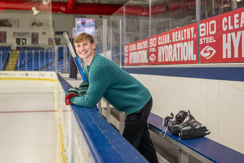 A male high school hockey player poses for a senior portrait standing in front of the bench at a hockey rink.  He is holding a hockey stick and wearing gloves