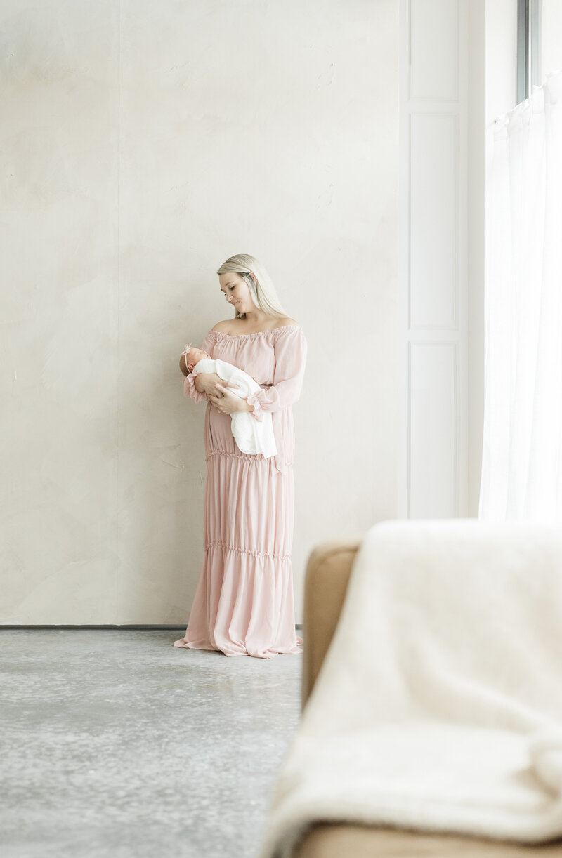 Mother in a soft pink gown standing against a neutral backdrop while gently holding her newborn, styled in a minimalist Kansas City newborn photography session.
